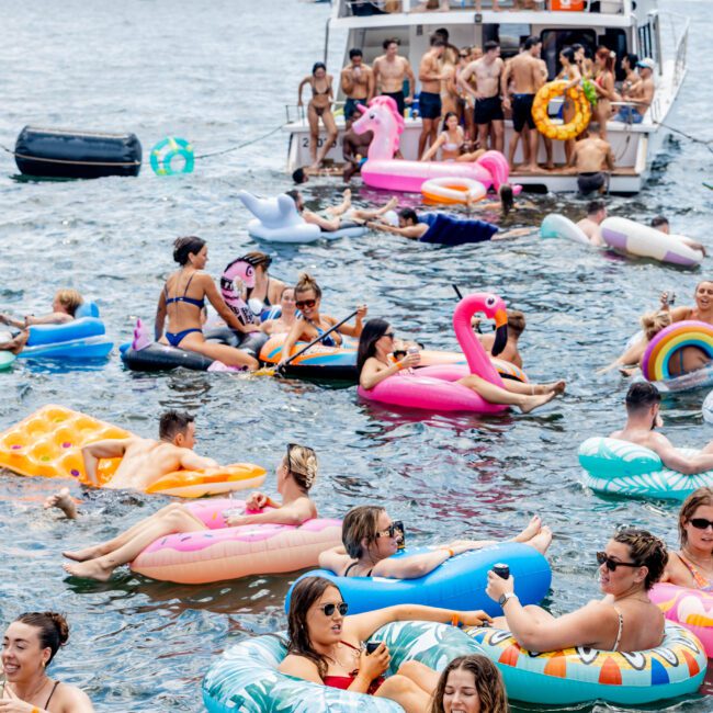 People are enjoying a sunny day on the water, lounging on colorful inflatables of various shapes like flamingos and unicorns. A boat is nearby with more people on board, and the background shows a distant shoreline and buildings.