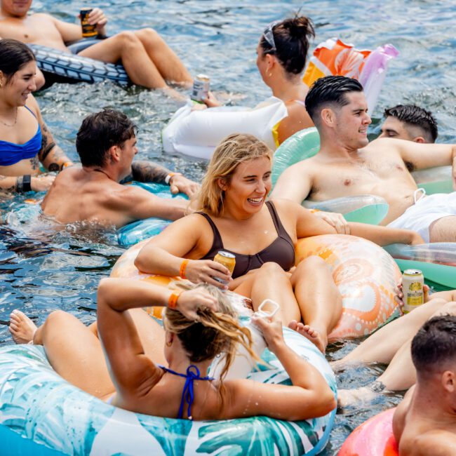 A group of people relaxing on colorful inflatable rings in a body of water, enjoying a sunny day. They are smiling and chatting, wearing swimwear. Some hold drinks, and various float designs are visible, including floral and solid colors.