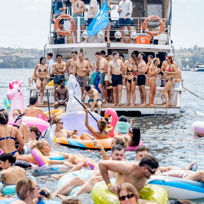 A large group of people on a boat and in the water enjoying a sunny day. The boat is crowded with individuals waving and smiling. In the water, numerous people are floating on colorful inflatables, including unicorns and donuts.