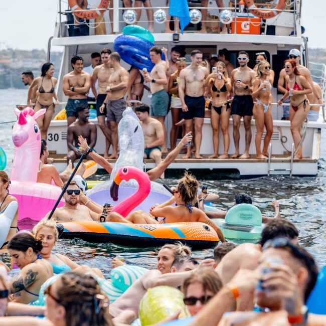 People enjoy a lively yacht party, with several floating in the water on inflatable toys shaped like flamingos and unicorns. Others are gathered on the boat, some dancing and taking photos, all under clear skies.