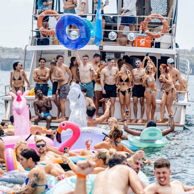 A lively boat party scene with people in swimsuits on a yacht and in the water. The crowd is enjoying summer with colorful inflatables like swans and flamingos. The atmosphere is festive and energetic under a clear sky.