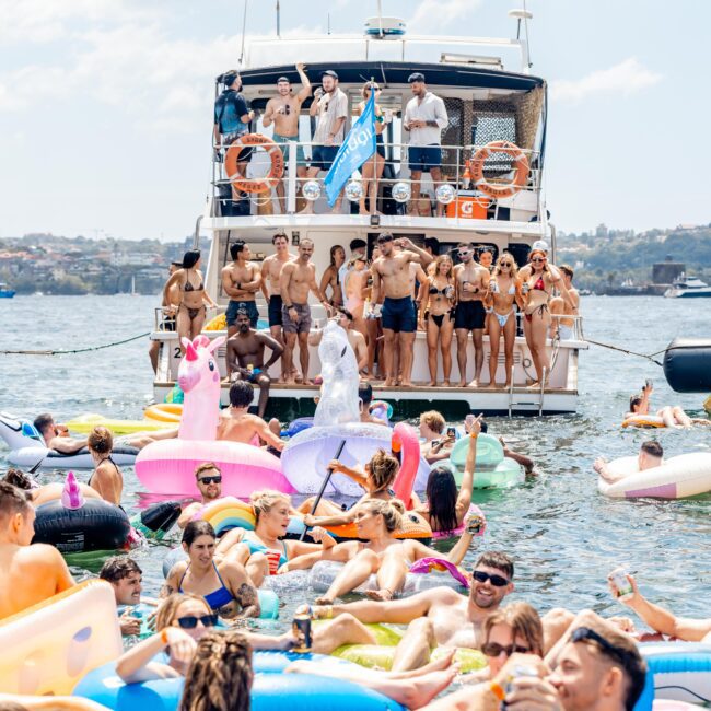 A lively scene on the water with many people on inflatable floats surrounding a boat. The boat, packed with people in swimwear, features a blue flag. The atmosphere is festive with clear skies and distant land visible.