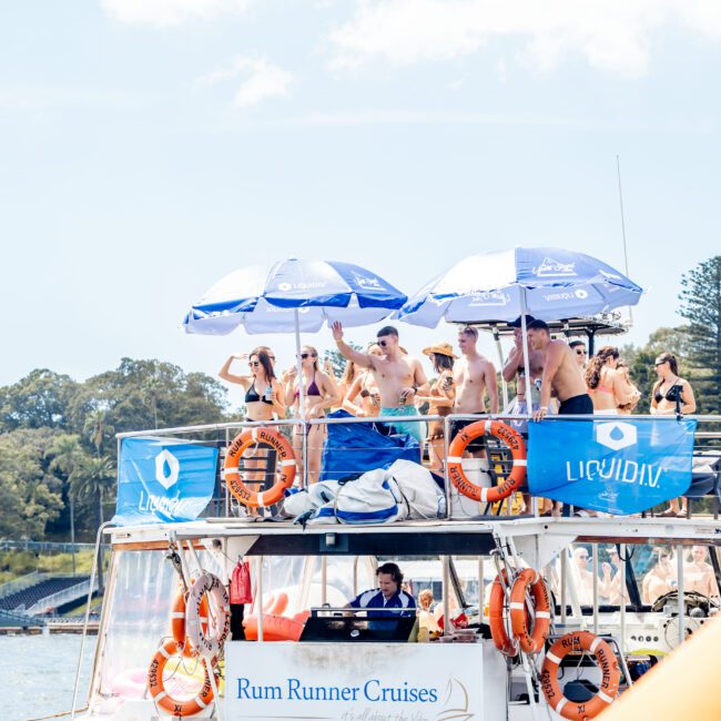 A group of people enjoying a sunny day on a double-decker boat labeled "Rum Runner Cruises." It has umbrellas on the top deck. The scene includes life rings, and trees in the background.