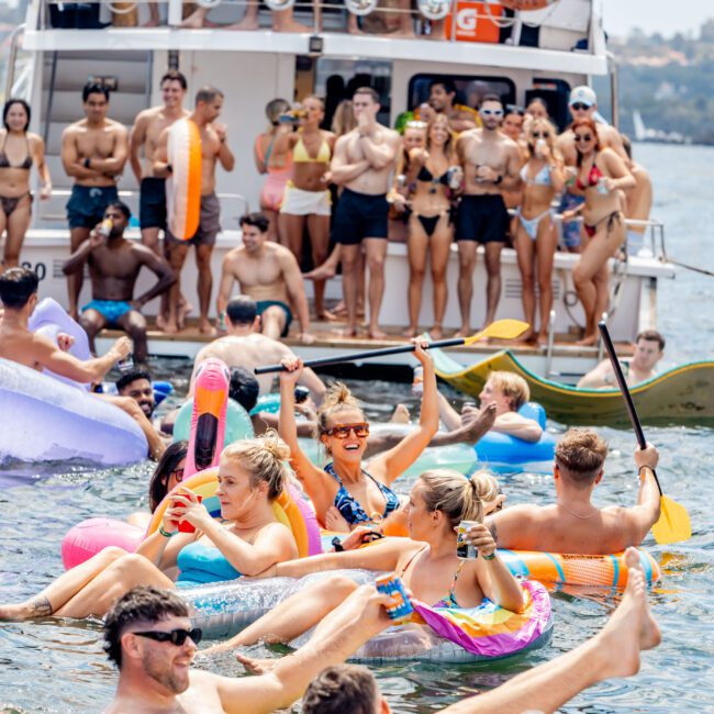 A lively scene of people enjoying a party on the water. Several individuals are floating on colorful inflatables in the water, while others are dancing on the deck of a boat in the background. The atmosphere is festive and carefree.