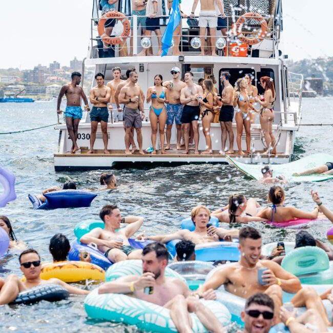 A group of people in swimsuits stands on the deck of a yacht, holding a flag. In the water, more people on inflatable floats enjoy a sunny day. The scene is lively and festive, with a city skyline in the background.