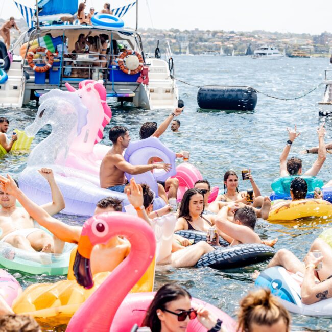 People enjoying a sunny day on the water with colorful inflatable floats, including a unicorn and flamingo. Two boats are docked nearby, with others socializing on deck, creating a lively and joyful atmosphere.