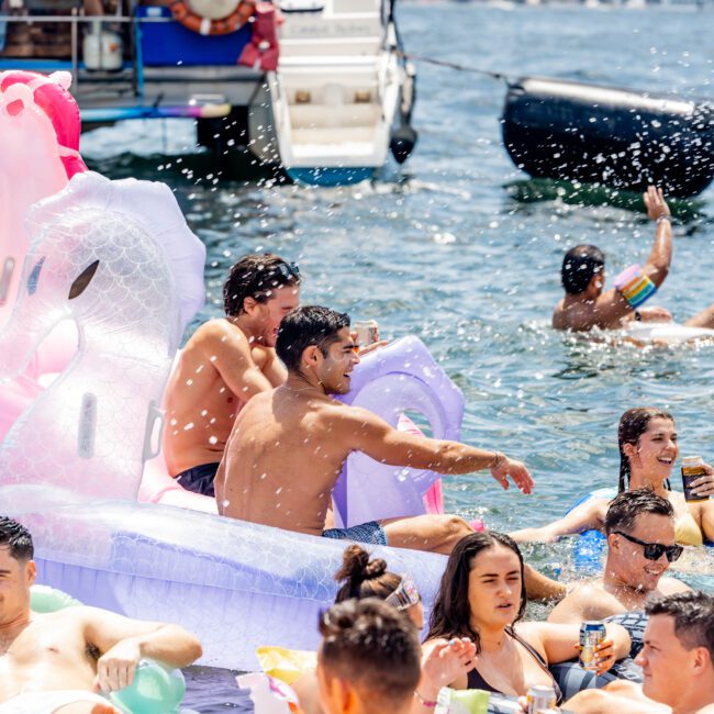 A group of people relax on inflatable floats in the water, enjoying a sunny day. A boat is docked nearby with more people on board. Some hold drinks, and water splashes in the air, creating a lively atmosphere.