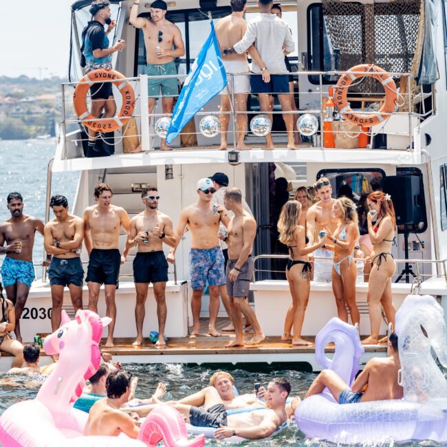 A group of people enjoy a sunny day on a boat and in the water. Some are on inflatable floats shaped like unicorns. Many are standing on the boat's deck, wearing swimwear, holding drinks, and socializing. A blue flag is visible on the boat.