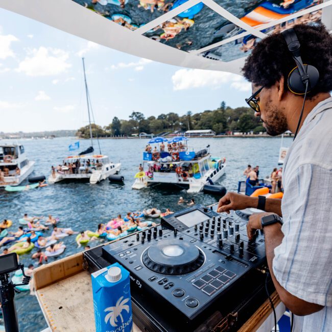 DJ wearing headphones plays music on a yacht deck with a mixer. Below, people in colorful inflatables enjoy a party in the water. Surrounding boats float nearby under a partly cloudy sky.