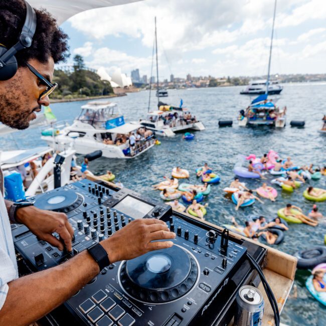 A DJ with headphones performs on a boat deck, overlooking a party on the water. People relax on colorful inflatables, surrounded by boats. The sea and distant shoreline are visible under a partly cloudy sky.