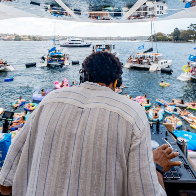 A DJ wearing headphones plays music on a boat surrounded by people floating on colorful inflatables in a lake. Other boats with partygoers are nearby, while a bright sky and cityscape appear in the background.