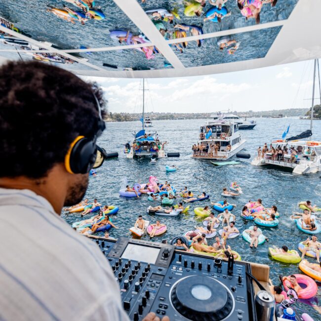 A DJ wearing headphones plays music on a boat while overlooking a lake party. People on colorful inflatables and nearby boats enjoy the scene under a sunny sky. Reflections are visible on a mirrored ceiling above the DJ.