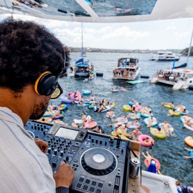 A DJ wearing headphones plays music on a boat deck, overlooking a lively scene with people on colorful inflatables in the water. Boats are anchored nearby under a blue sky. A reflective surface shows the crowd.