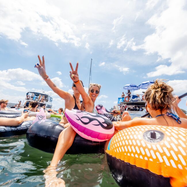 People relaxing on inflatable tubes in a lake, surrounded by boats. A person in the center has their arms raised, flashing peace signs. The sky is blue with some clouds, creating a lively, festive atmosphere.