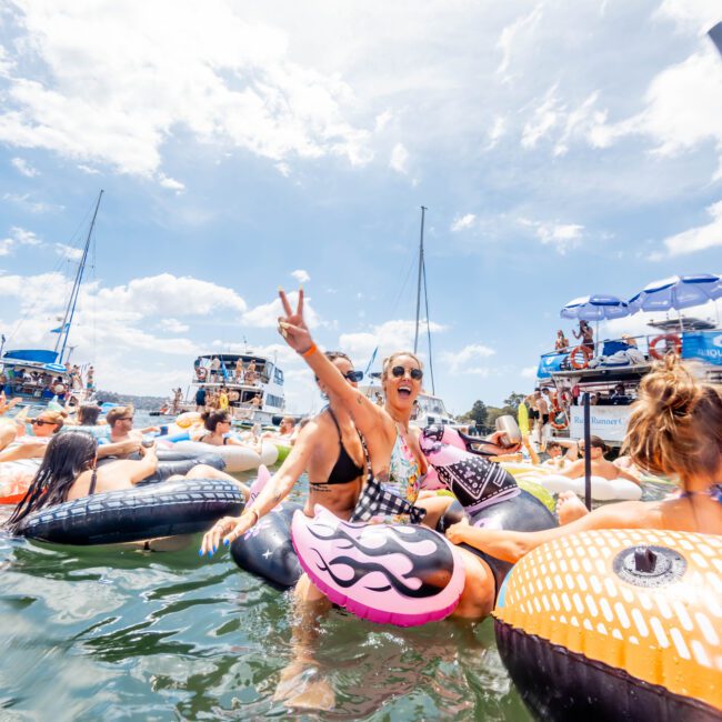 A group of people enjoying a sunny day on the water, sitting on inflatable rafts. The main person is wearing sunglasses and making a peace sign. Boats and other people are in the background, with a partly cloudy sky above.