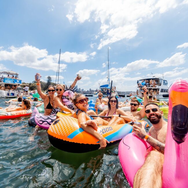 A group of people enjoying a sunny day on colorful inflatable floaties in the water. They are smiling, holding beverages, and surrounded by more floats and boats in the background. The sky is clear with a few clouds.