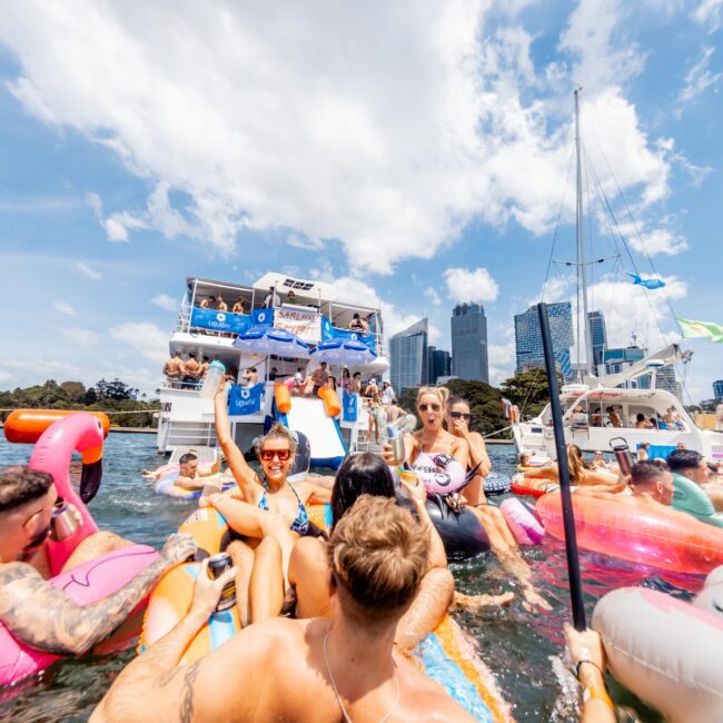 A lively gathering of people on colorful inflatable floats in a sunny harbor. A large yacht with a party visibly ongoing is in the background, set against a city skyline with tall buildings. The sky is blue with scattered clouds.