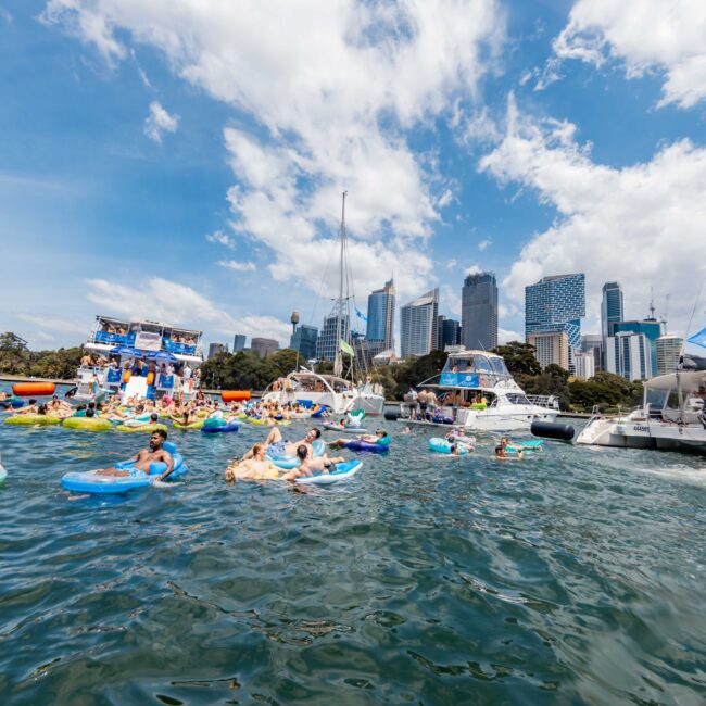 People are enjoying a sunny day on the water, floating on colorful inflatables in a harbor. Several yachts are anchored nearby, and tall city buildings are visible in the background under a partly cloudy sky.