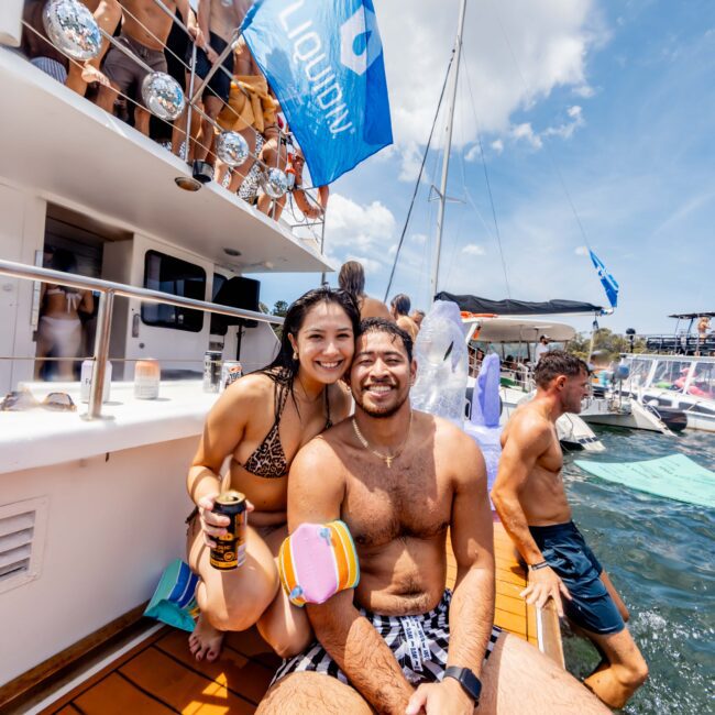 A man and woman in swimwear sit together on a boat's deck, smiling at the camera. The woman holds a drink, and people are visible in the background on the boat. A blue flag with white writing is flying in the breeze. The water and sky are clear and bright.