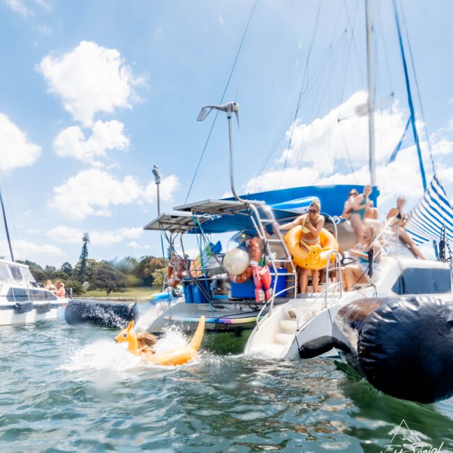 A group of people on a yacht enjoying a sunny day. A person is sliding into the water on a kangaroo-shaped inflatable. The yacht is anchored near the shore, surrounded by other boats. The sky is clear with a few clouds.