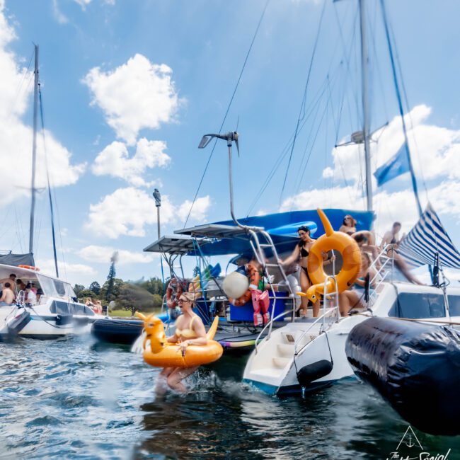 People enjoying a sunny day on boats, with some in the water using inflatable pool floats like a unicorn and a duck. The water is clear, and the sky is partly cloudy.