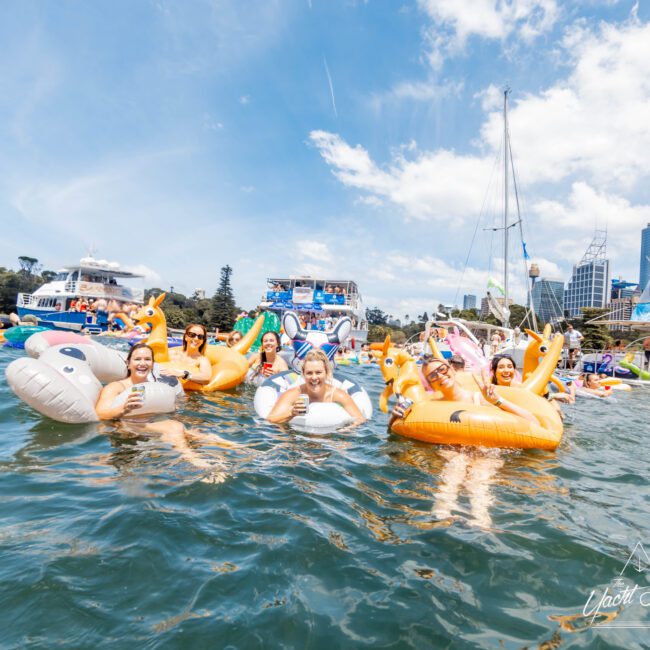 A group of people enjoying the sunny day on colorful inflatable rafts in the water. In the background, several boats are anchored. The scene is lively, with clear blue skies and high-rise buildings visible in the distance.