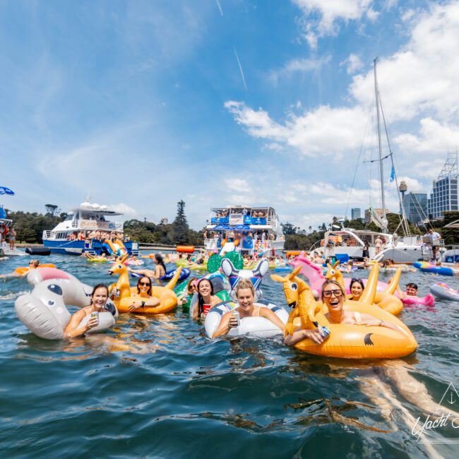 A group of people smiling and relaxing on inflatable floats shaped like unicorns and other animals in a sunny harbor. Several boats are anchored nearby, and city buildings are visible in the background. The sky is clear, indicating a bright day.