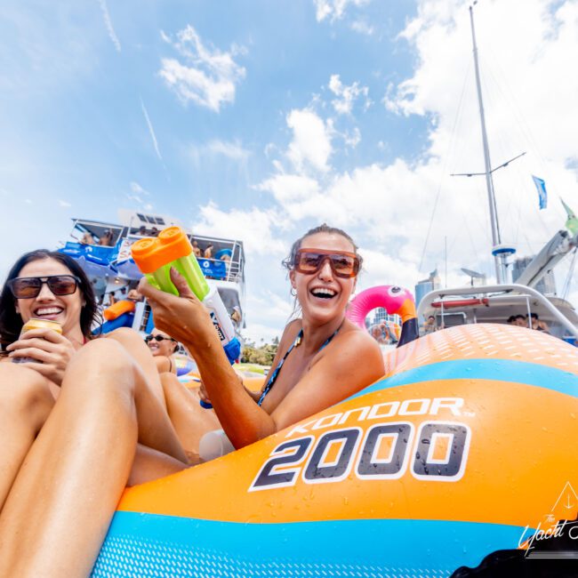 A group of people laughing and relaxing on a colorful inflatable raft in the water. The sky is clear and blue, with surrounding boats in the background. One woman holds a drink container, and everyone appears to be enjoying a sunny day.