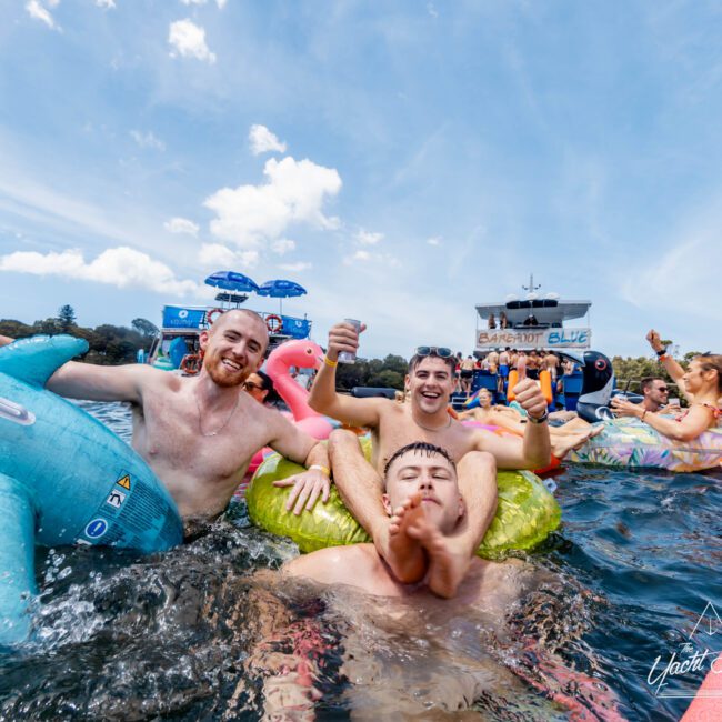 A group of people are having fun in the water, sitting on colorful inflatable floats. They're laughing and giving thumbs up. In the background, there's a boat with a banner that says "Yacht Social." The weather is sunny with some clouds.