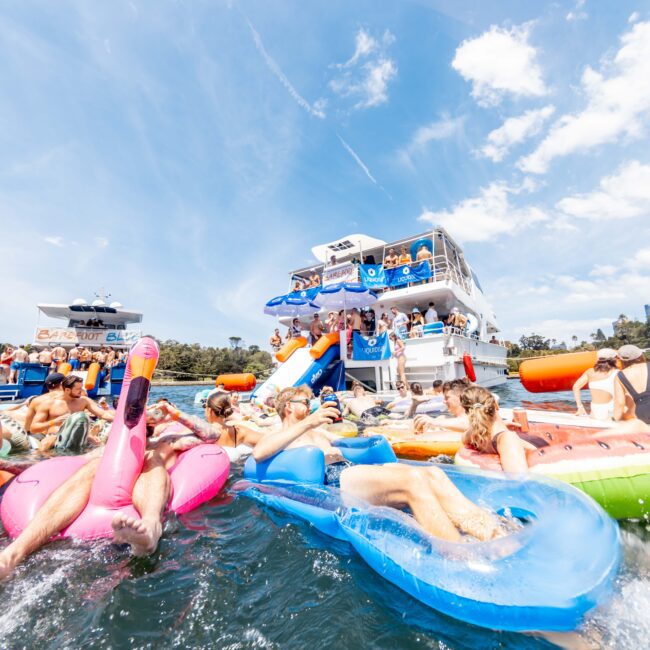 A group of people enjoy a sunny day on the water, lounging on colorful inflatable floats near a boat. The sky is blue with scattered clouds. People are swimming and relaxing in a festive, joyful atmosphere.