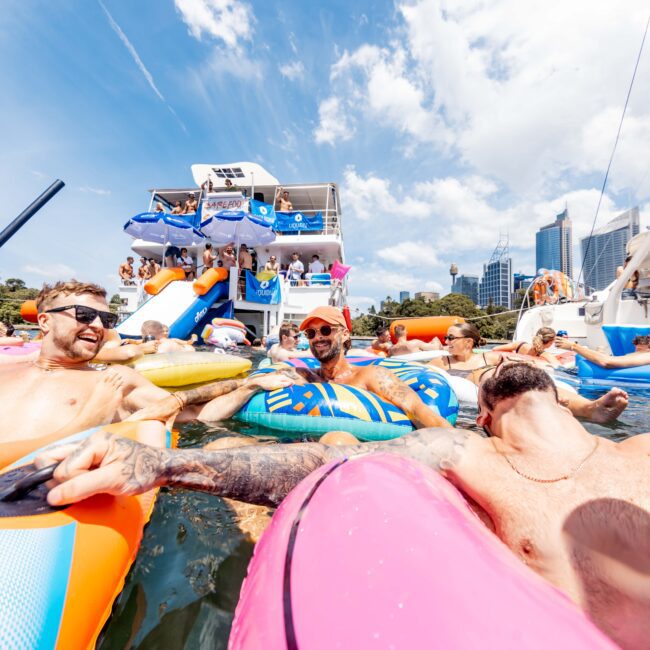 A lively scene of people enjoying a pool party on colorful inflatables in the water. A docked boat with a slide is filled with more party-goers. It's a sunny day with a bright sky and cityscape in the background.