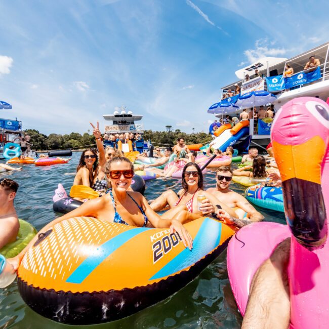 A group of people enjoying a day on the water, lounging on colorful inflatable floats including a pink flamingo. The sunny sky and a party boat in the background create a festive atmosphere. Participants are smiling, wearing swimsuits and sunglasses.