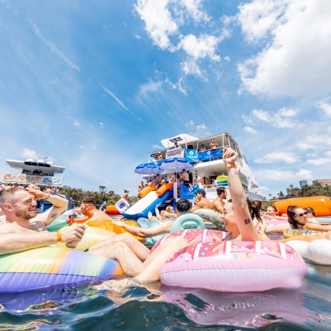 People relaxing on colorful inflatable pool floats in sunny outdoor water. A crowd gathers near a floating bar with a sign reading "Back to the Bay." Blue skies and fluffy clouds in the background enhance the festive atmosphere.