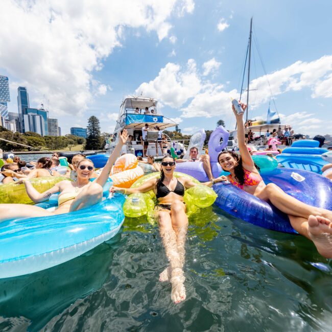 A group of people are relaxing on colorful inflatable floats in the water. They are smiling and posing for the camera. In the background, boats and a city skyline are visible under a partly cloudy sky.