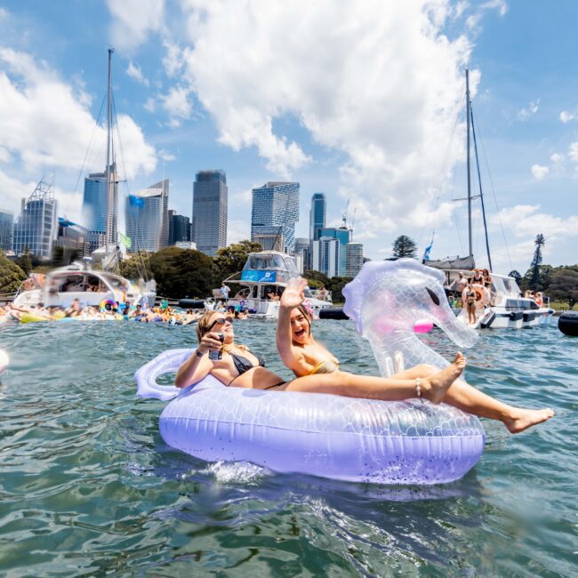 Two people relax on a pink inflatable pool float in the water, with a city skyline and boats in the background. They smile and wave, wearing swimsuits under clear skies.