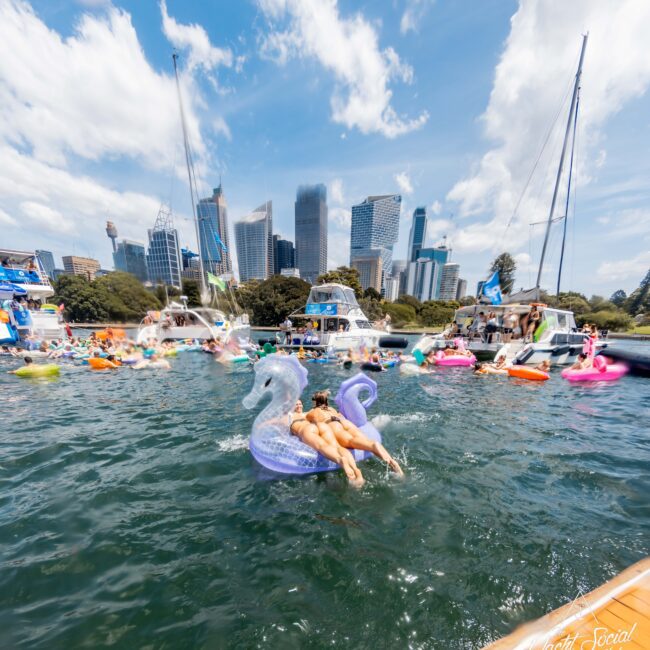 People enjoying a sunny day on inflatable floats in a city harbor, with skyscrapers in the background. A person is lounging on a seahorse-shaped float in the foreground. Bright blue sky with scattered clouds.