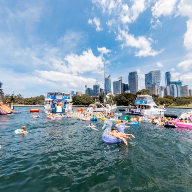 People on colorful inflatables enjoy a sunny day in the water near several boats. Skyscrapers and trees are visible in the background under a bright blue sky.