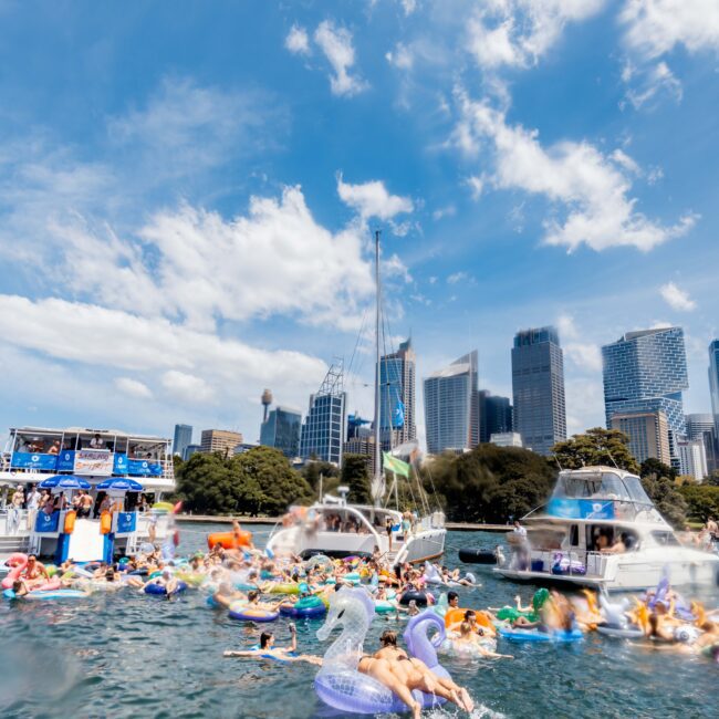 A sunny outdoor scene on the water with multiple people relaxing on colorful inflatable tubes. Boats are anchored nearby, and a city skyline with skyscrapers and greenery is visible in the background under a blue sky with clouds.