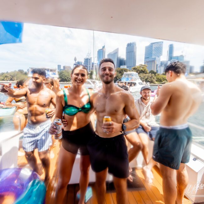 A group of people enjoying a sunny day on a boat. Two people pose in the foreground holding drinks, surrounded by others socializing. Skyscrapers and trees are visible in the background under a partly cloudy sky.