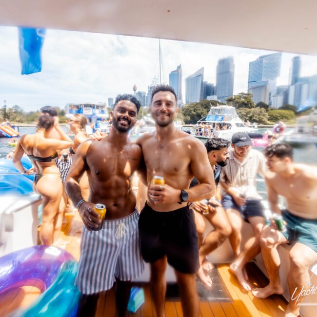 Two men pose on a yacht, each holding drinks. People in swimsuits are relaxing in the background. The scene is lively with a city skyline visible in the distance. It’s a bright day.