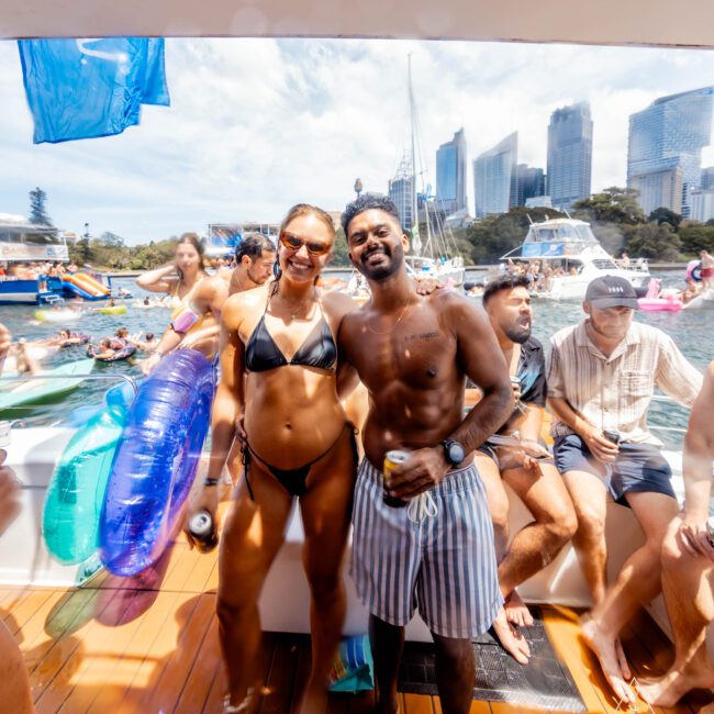 A group of people enjoying a sunny day on a boat. A woman in a black bikini and a man in striped shorts are smiling at the camera. The boat is crowded and there are skyscrapers and other boats in the background.