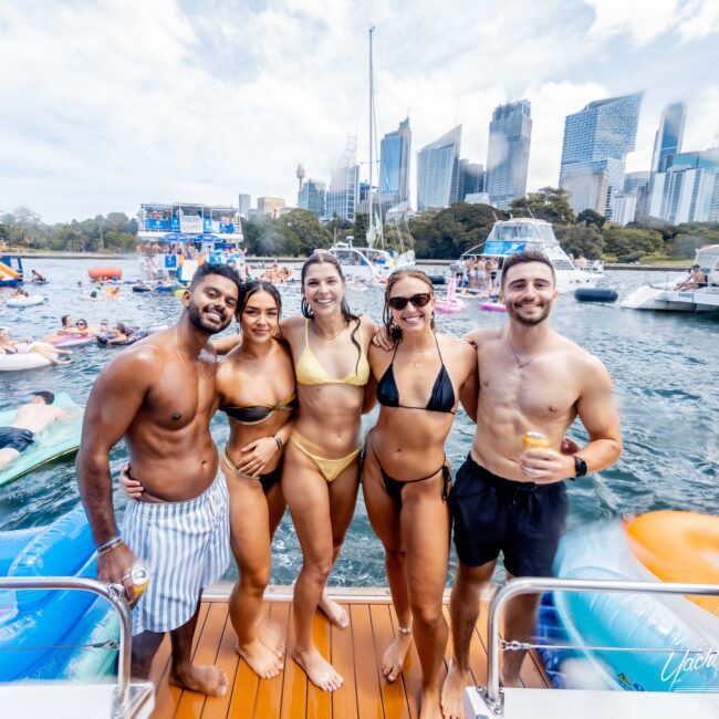 A group of five people in swimsuits stand and smile on a boat deck. The background shows a marina with other boats, inflatables, and a city skyline under a partly cloudy sky.