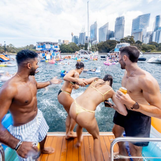 A group of people on a boat, with two individuals playfully pushing a woman in a yellow bikini into the water. Other boats and people are in the background, and city buildings are visible under a partly cloudy sky.