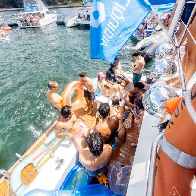 A group of people in swimsuits gather on the deck of a boat under a blue flag on a sunny day. The boat is docked on a body of water with other boats visible in the background. Trees line the distant shore.