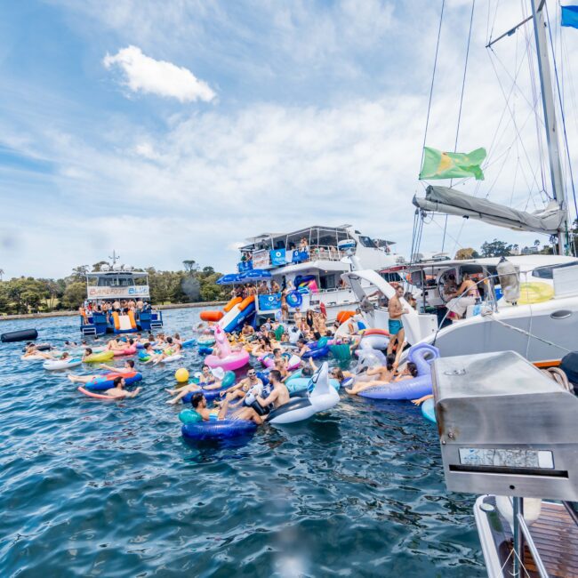 A lively gathering of people enjoying a sunny day on a lake. Many are lounging on colorful inflatables shaped like animals and boats. Several yachts and boats are nearby, with green trees and a blue sky in the background.