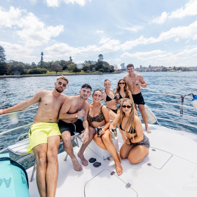 A group of six people sit and pose on a boat, smiling under a sunny sky. They are dressed in swimwear and holding drinks. The backdrop features water and a distant shoreline with trees and a few buildings.