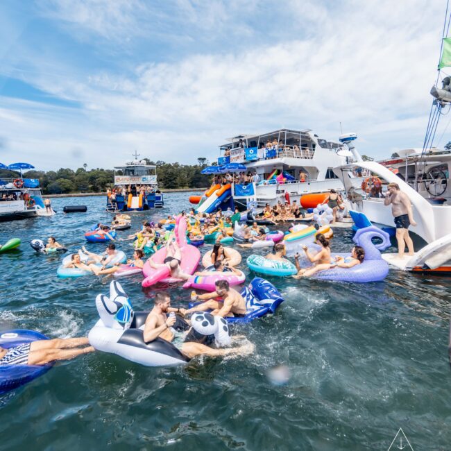 A lively gathering on the water features people on inflatable floats shaped like animals and unicorns. Several boats are anchored nearby, with more people onboard. The sky is partly cloudy, and the scene conveys a festive, summer atmosphere.