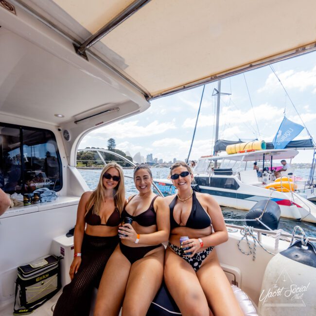 Three people in swimsuits sit smiling on a boat, holding drinks. They are under a canopy with another boat visible in the sunny background. The scene suggests a lively and relaxed day on the water.