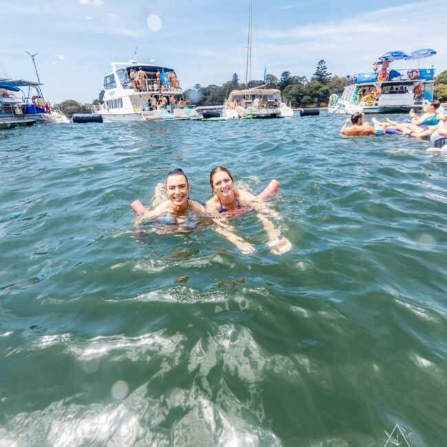 Two people swim in clear green water with several boats and people in the background. The sky is blue with few clouds. Logs and structures are visible in the distance. The image has a watermark in the bottom right corner.