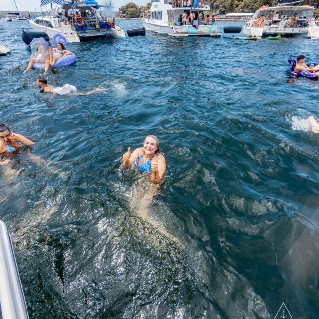 A person swimming in the ocean near several anchored boats. People are lounging on the boats and in the water. The swimmer smiles and gives a thumbs-up. The scene is sunny and festive, with clear skies above.